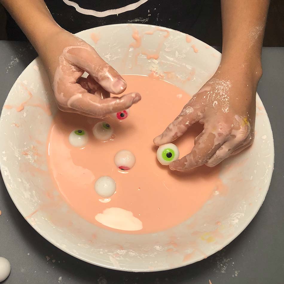 Des mains d'enfant manipulent des jouets en forme de globes oculaires dans un liquide gluant et rosâtre.
