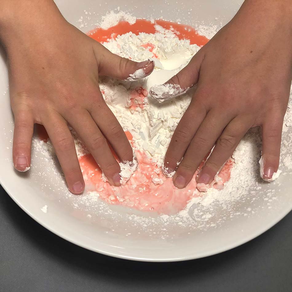 Gros plan de mains d'enfant mélangeant un liquide rouge à de l'amidon de maïs dans un plat.