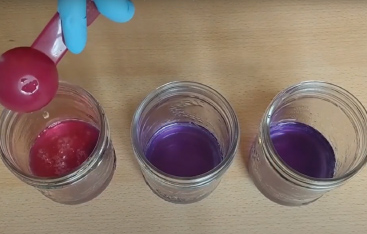 A gloved hand dumps liquid from a spoon into a jar containing a red liquid, sitting next to two jars containing a purple liquid.