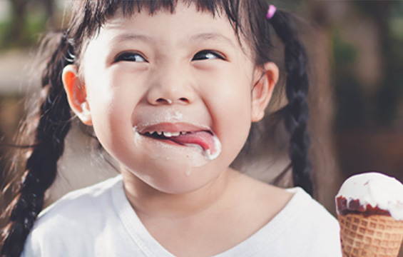 A young girl holding an ice cream cone licks ice cream from her face.