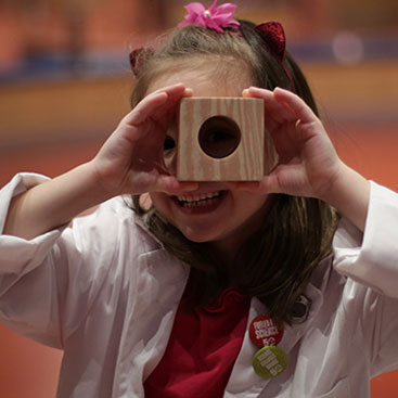 Une fille en sarrau regarde au travers du trou d'un cube de bois.