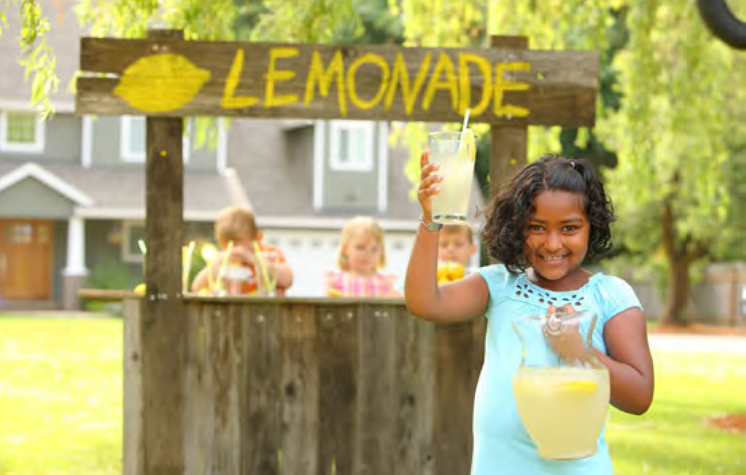 Une fille tient un pichet et un verre de limonade devant un comptoir de vente de limonade.