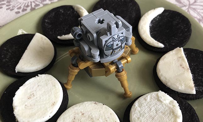 Sandwich cookies are laid out on a plate in a circle, with their cream centres carved away to look like different phases of the moon. A toy moon lander sits in the middle of the cookies.