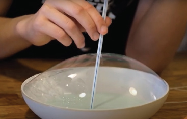 A straw is inserted into the middle of a large bubble that is sitting in a shallow bowl.
