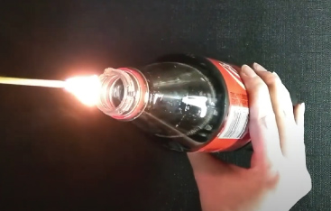 A lit match is held next to the opening of a full soda pop bottle.
