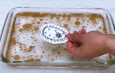 A hand holds a paper model of a microbe above a pan filled with water and a brown oil.