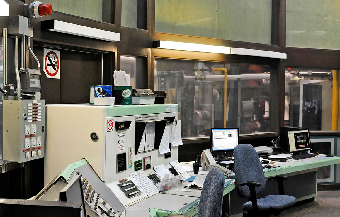 Une salle de contrôle où se trouvent des ordinateurs avec des machines derrière une baie vitrée.