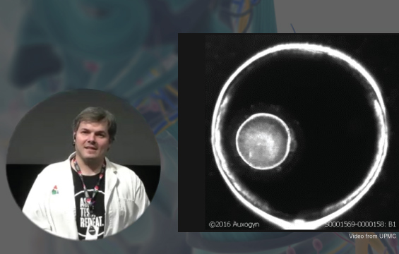 Image d'une cellule en division sous un microscope; un éducateur paraît dans une fenêtre au coin gauche.