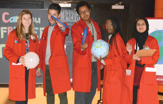 Cinq élèves de l'École des sciences vêtus de leur sarrau rouge tenant chacun un objet, deux tenant un ballons, deux tenant un Frisbee, une tenant un presse-papier.