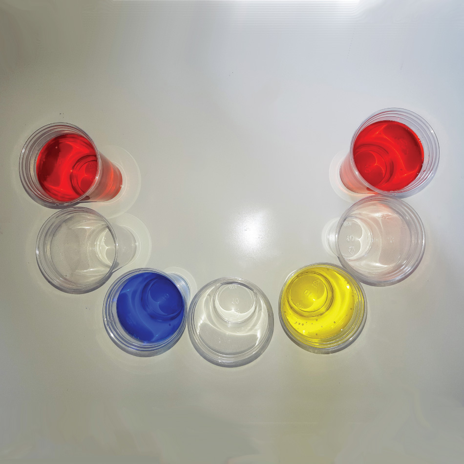 Sept verres en plastique disposés en demi-cercle, remplis d'eau colorée ou non, de gauche à droite dans l'ordre suivant : rouge, limpide, bleu, limpide, jaune, limpide, rouge.