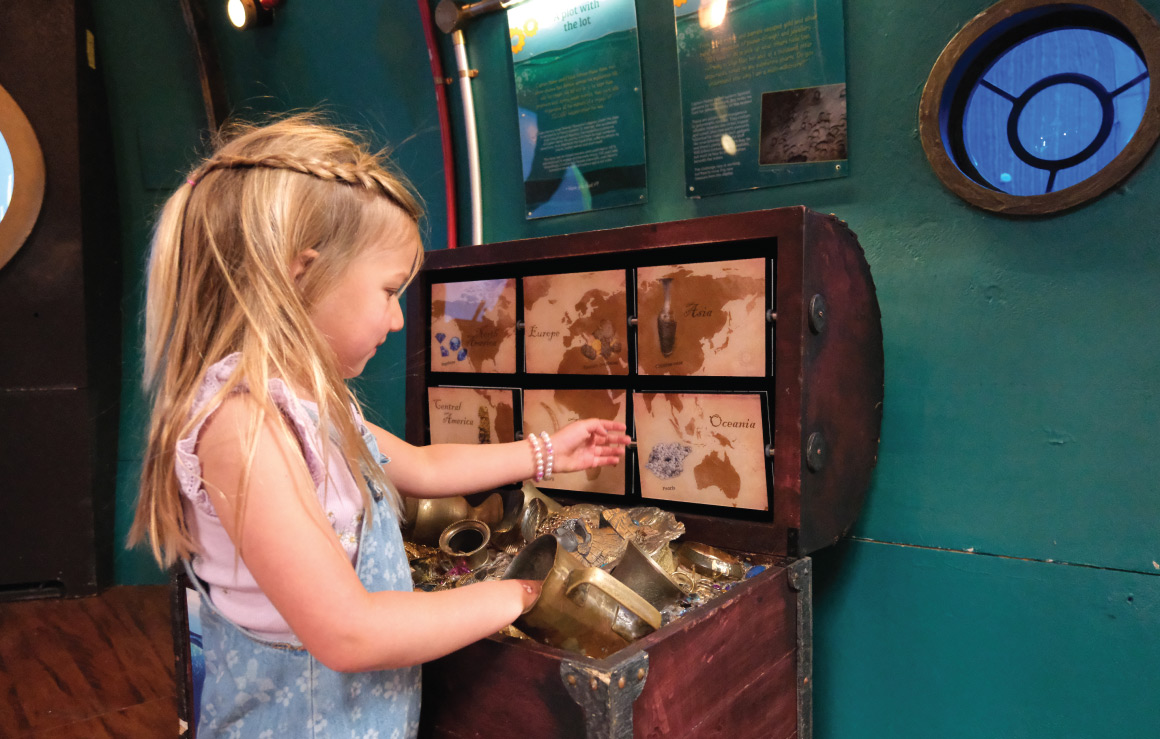 Une enfant fouille dans un coffre rempli d'objets dorés.