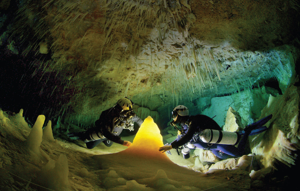 Deux personnes munies d'équipement de plongée touchent une stalagmite dans une grotte sous-marine.