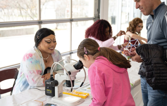 Fierté dans les STIM 2024 : une fille regarde au microscope d'une drag queen en sarrau multicolore.