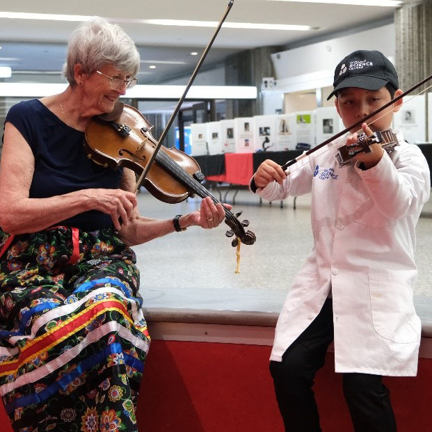 Une femme âgée en jupe métisse et un garçon en sarrau jouent du violon ensemble / An elderly woman in a colourful skirt and a boy in a lab coat play the violin together