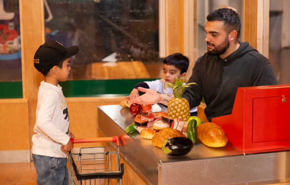 Un père et ses deux fils jouent à la caisse avec des fruits et des légumes.