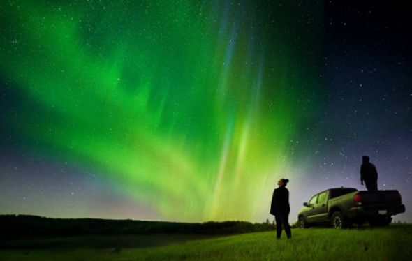 Two adults in a field with a pick-up truck are observing a green aurora borealis in a starry sky, one person standing on the truck and the other standing next to it.